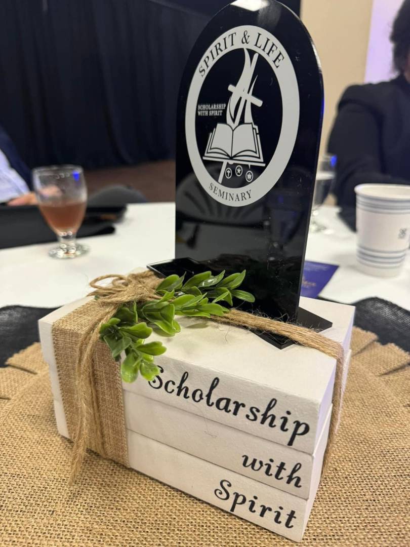 a decorative table centerpiece from an event, featuring a black, tombstone-shaped plaque with the logo and text "Spirit & Life Seminary" and "Scholarship with Spirit." The plaque is placed on top of a stack of three white, book-like blocks tied together with twine and decorated with a small green plant. The text "Scholarship with Spirit" is also written on the side of the book-like blocks.
