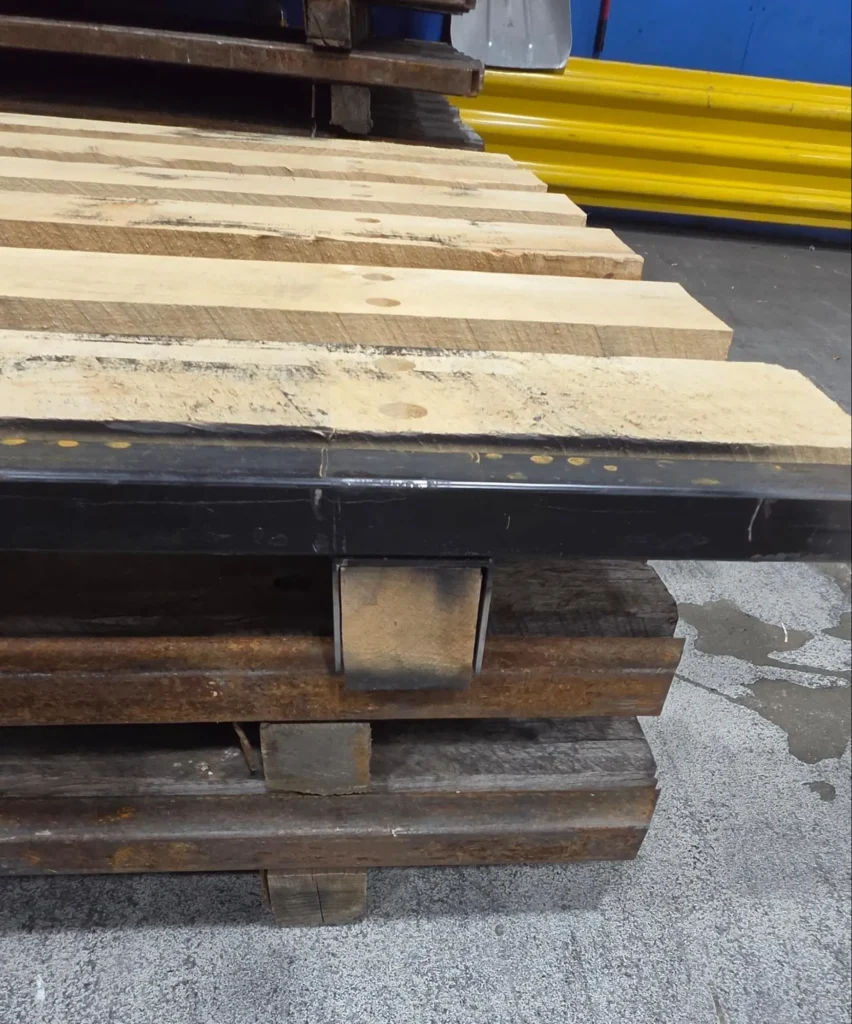 Close-up of a repaired steel-reinforced pallet with new wide hardwood lead boards and a reinforced fastening pattern mounted on a black steel frame, stacked on worn steel pallets waiting for repair.