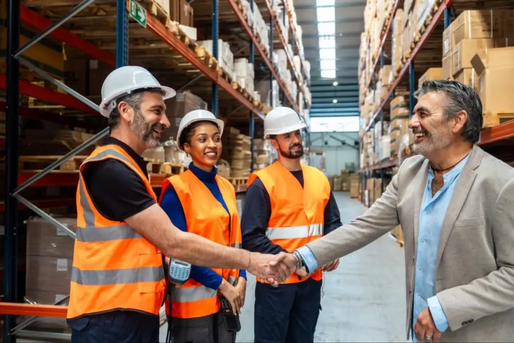 Two professionals shaking hands in a warehouse facility with pallets and logistics operations in the background, representing a strategic sourcing partnership.